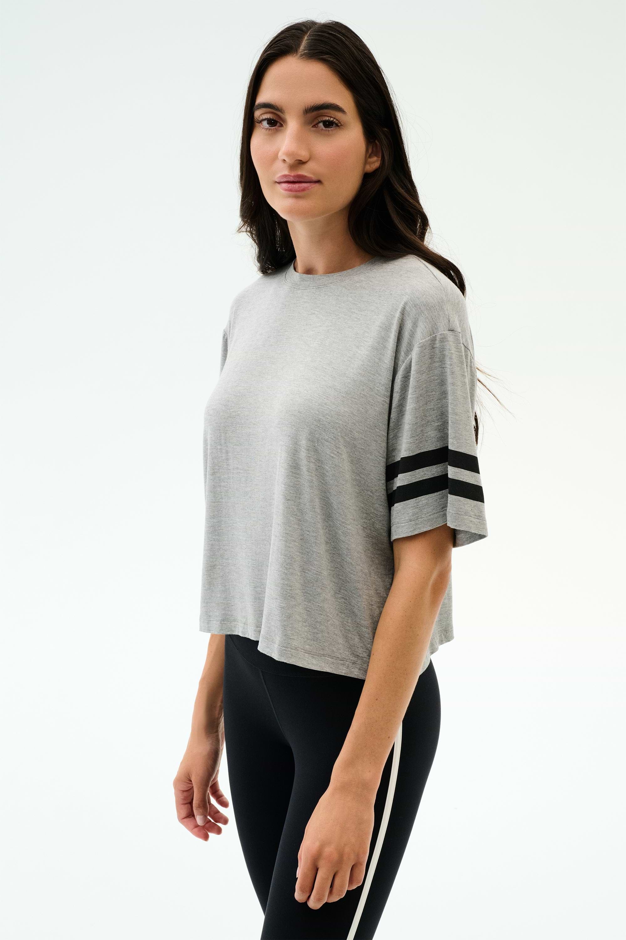 A woman stands against a plain background wearing the SPLITS59 Ava Jersey Tee in heather grey/black with black sleeve stripes and black leggings—ideal for running or intense gym workouts.