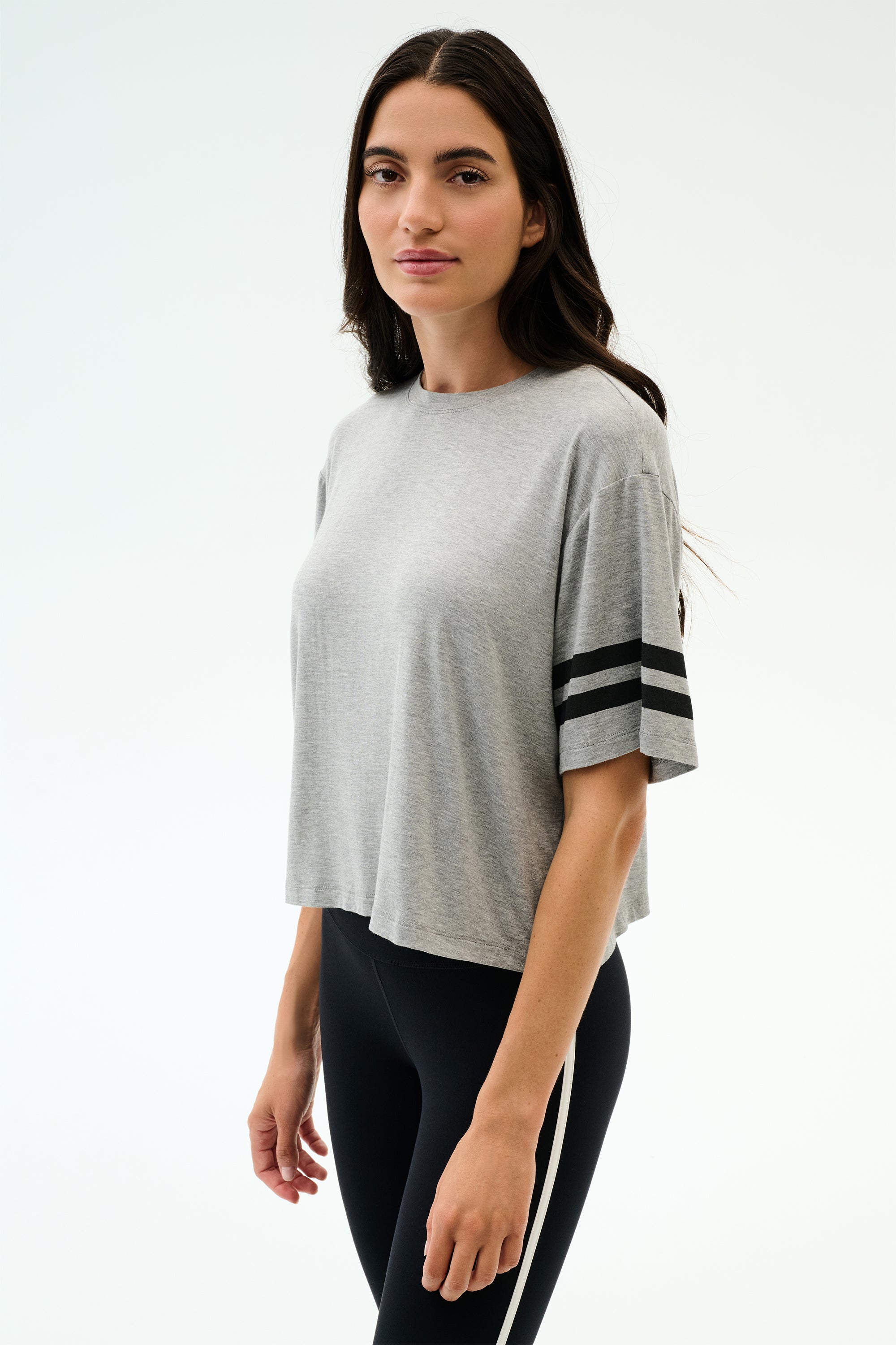 A woman stands against a plain background wearing the SPLITS59 Ava Jersey Tee in heather grey/black with black sleeve stripes and black leggings—ideal for running or intense gym workouts.