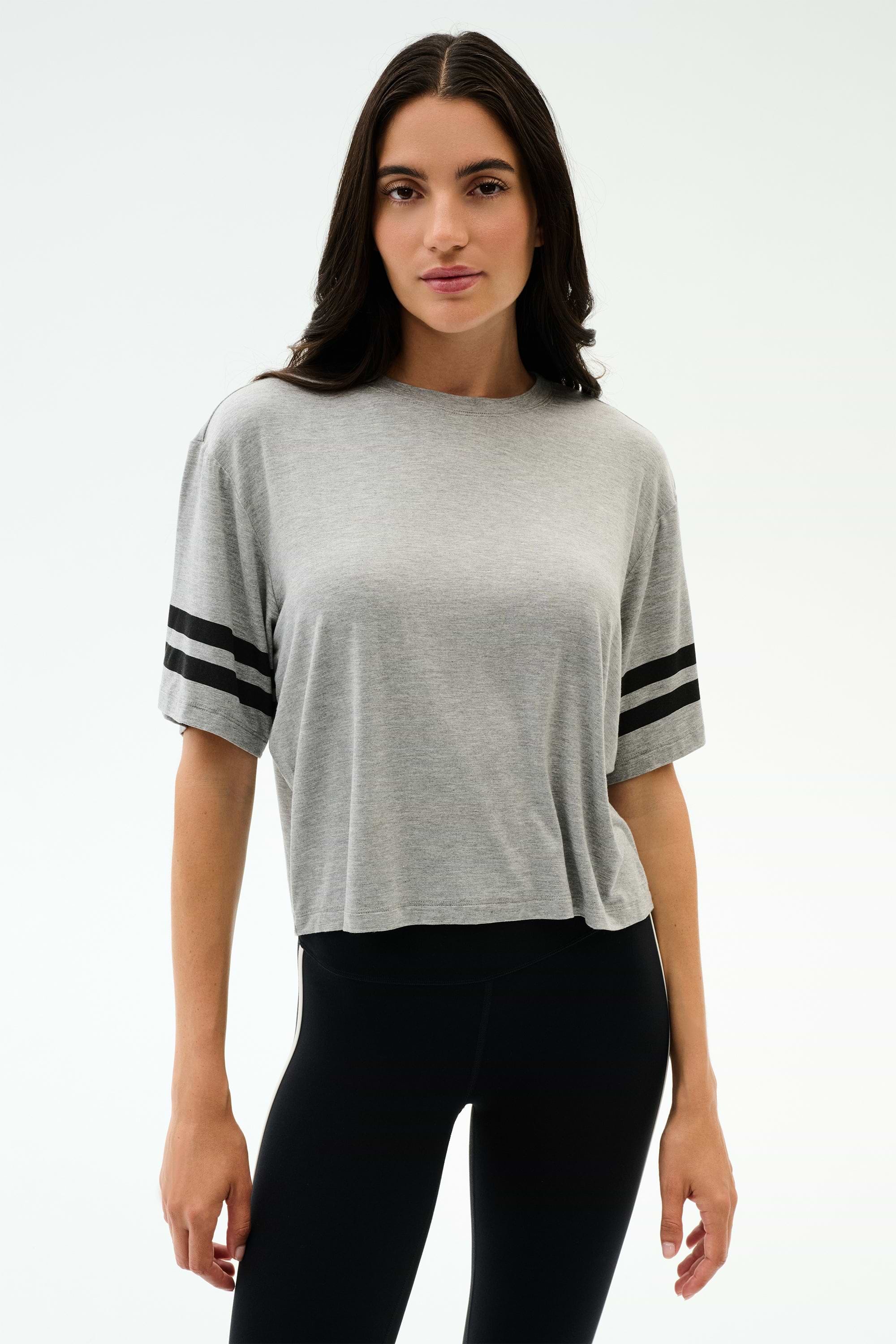 A person stands against a plain background wearing the SPLITS59 Ava Jersey Tee in Heather Grey/Black and black leggings, perfect for gym workouts or running.