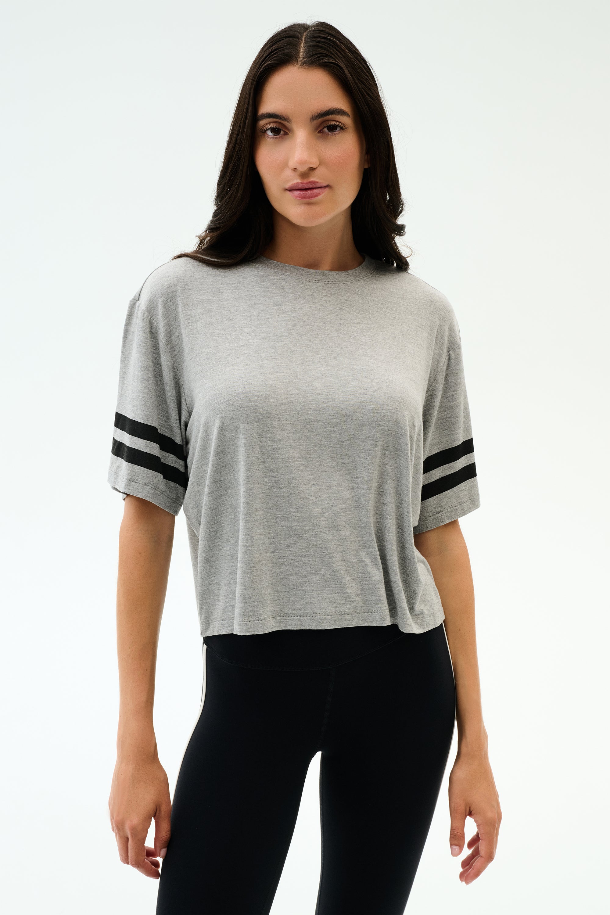 A person stands against a plain background wearing the SPLITS59 Ava Jersey Tee in Heather Grey/Black and black leggings, perfect for gym workouts or running.