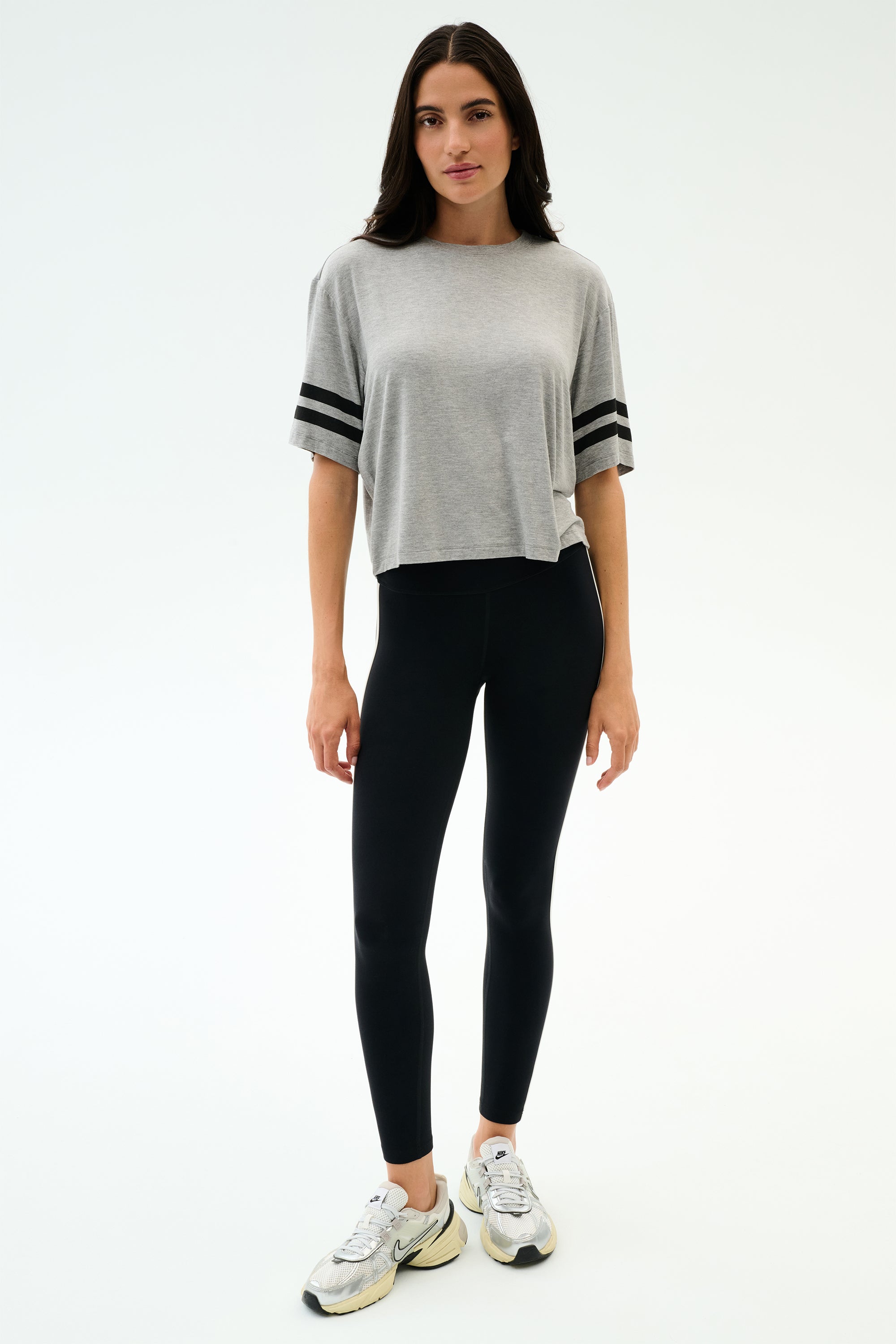 A woman poses against a white backdrop in the SPLITS59 Ava Jersey Tee - Heather Grey/Black, styled with black leggings and white athletic shoes—ideal for workouts or running.