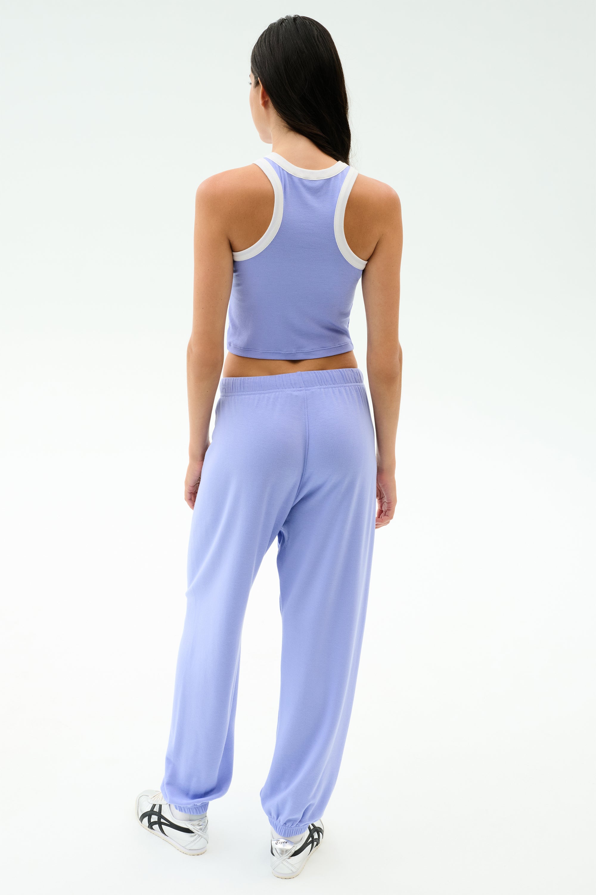 A woman stands facing away, wearing SPLITS59’s Andie Oversized Fleece Sweatpant in Purple Haze with a light purple tank top and white sneakers against a plain backdrop—perfect for warm weather workouts.