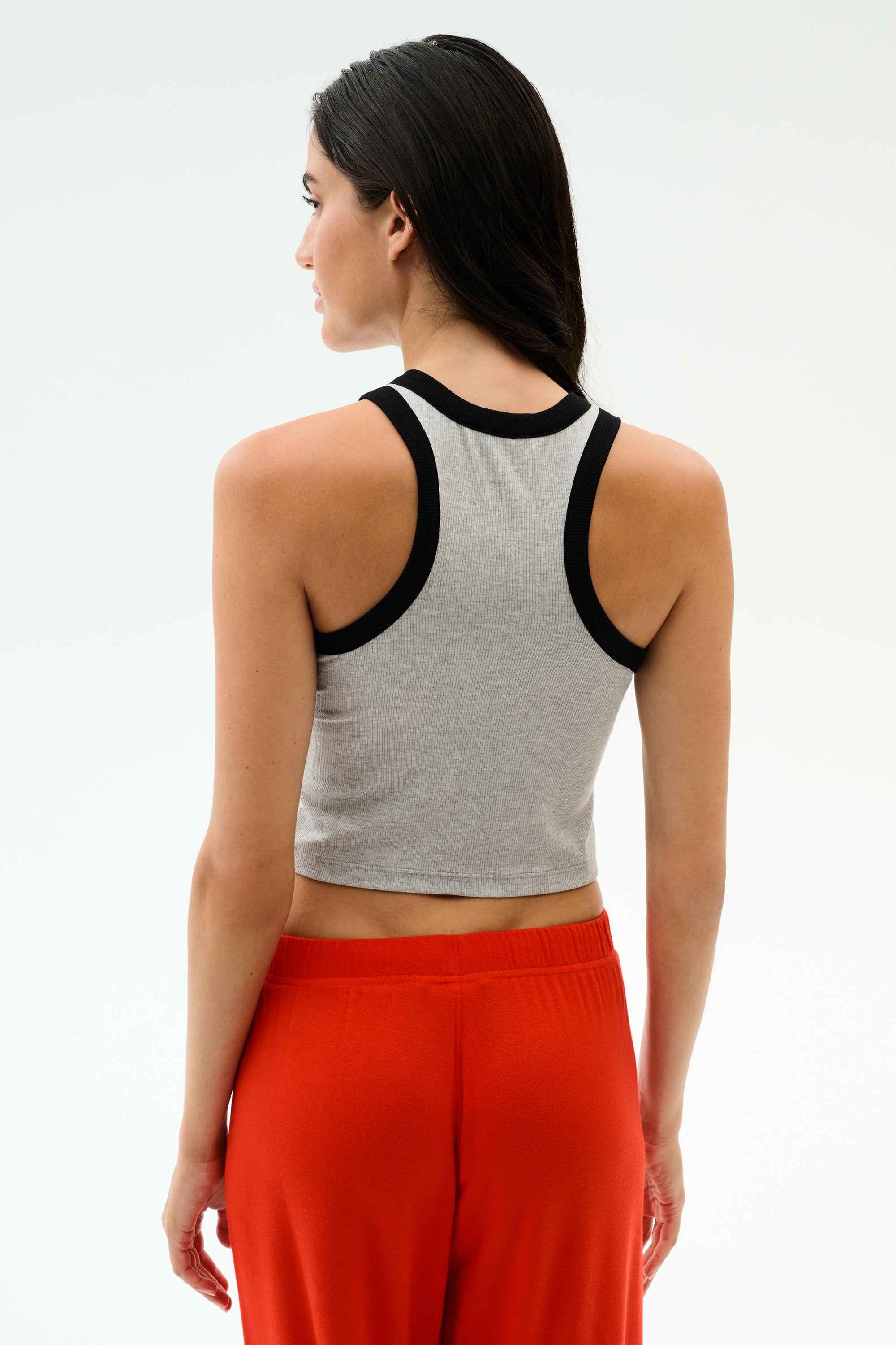 A woman faces away, wearing the SPLITS59 Kiki Rib Crop Tank in Heather Grey/Black, paired with bright red pants against a plain light background.
