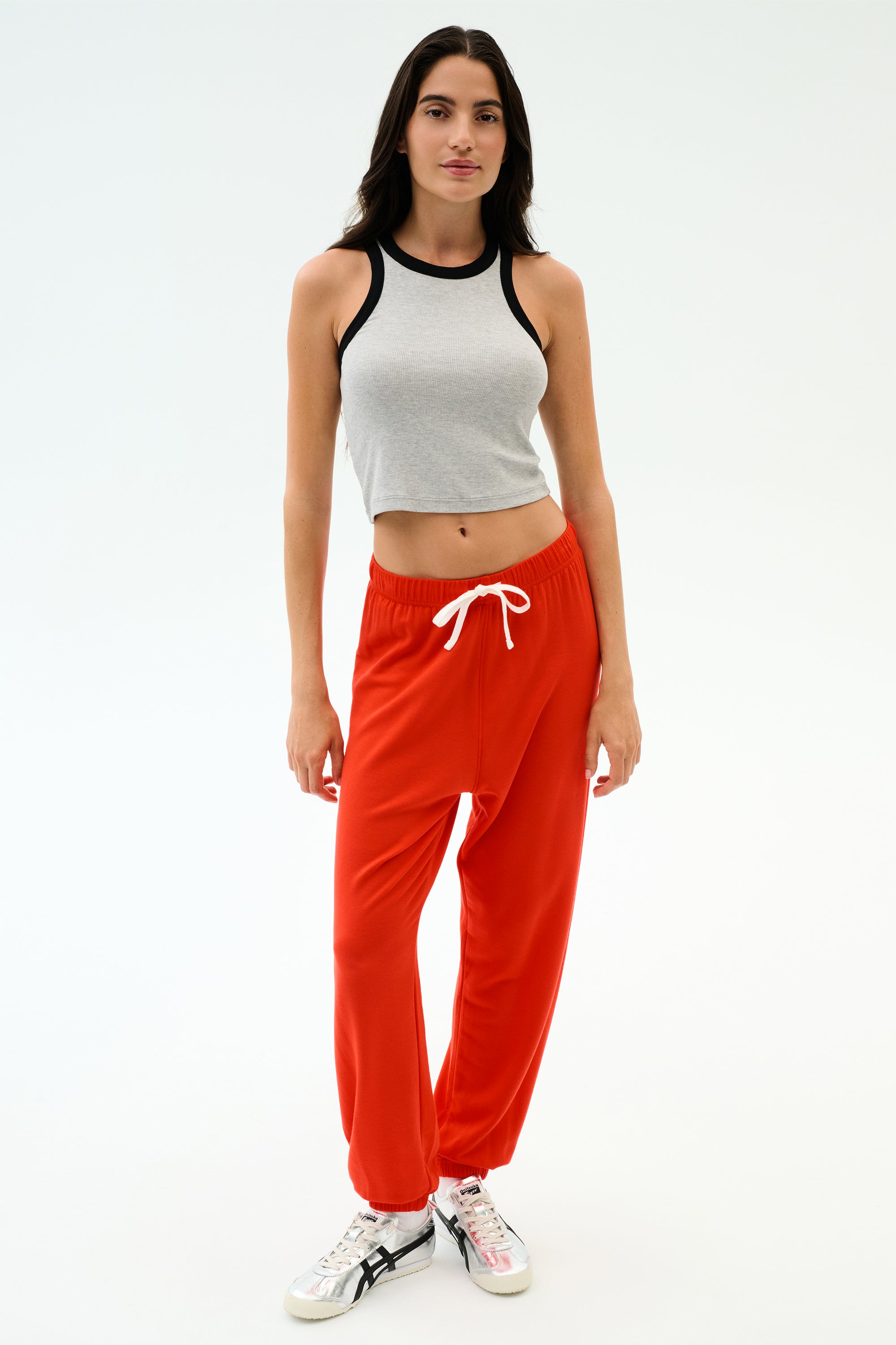A woman stands against a white background wearing the SPLITS59 Kiki Rib Crop Tank in Heather Grey/Black, red sweatpants with a white drawstring, and white athletic shoes.