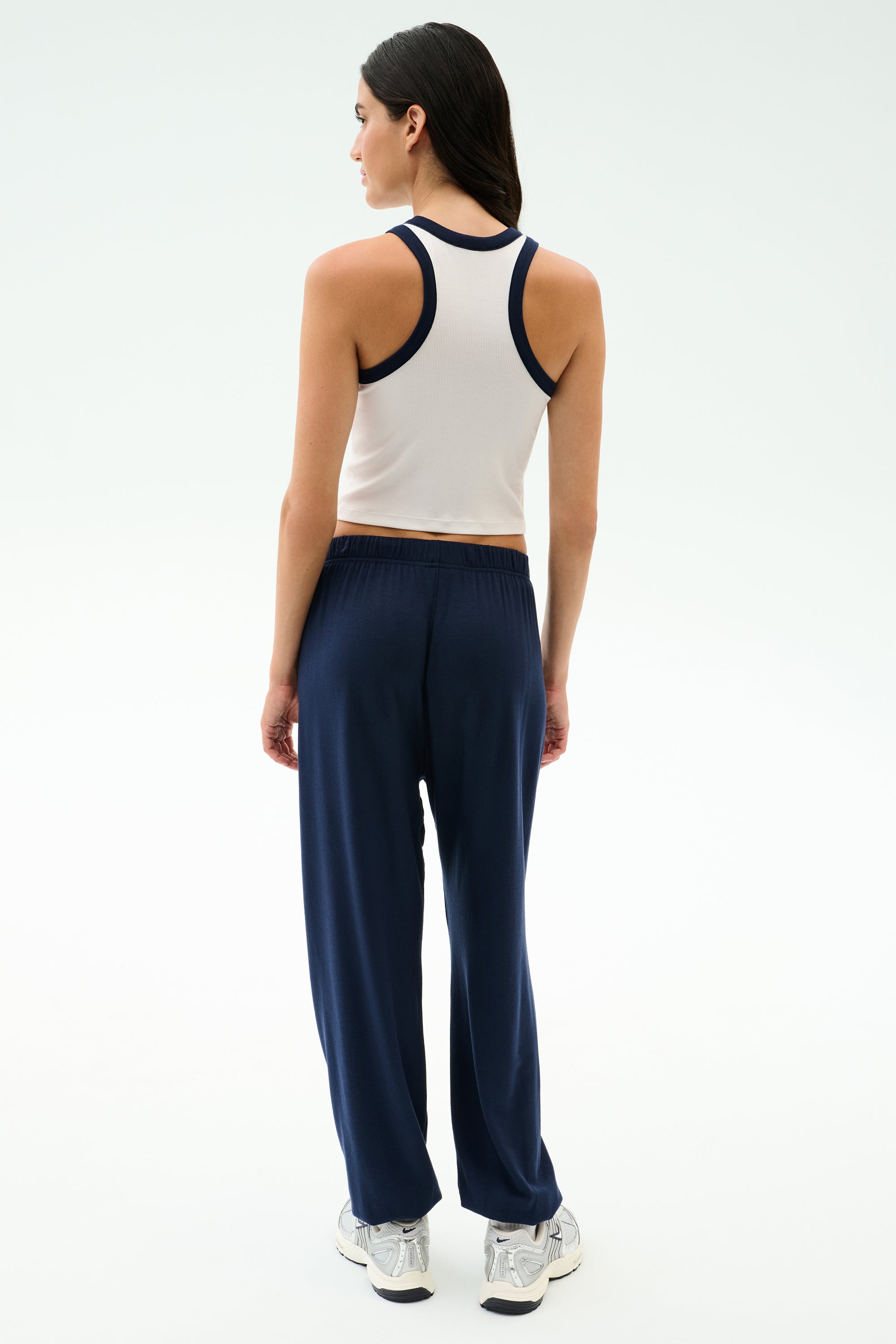 A woman faces away, wearing the SPLITS59 Kiki Rib Crop Tank in White/Indigo, paired with navy pants and white sneakers against a plain light background—ideal for gym workouts or casual style.