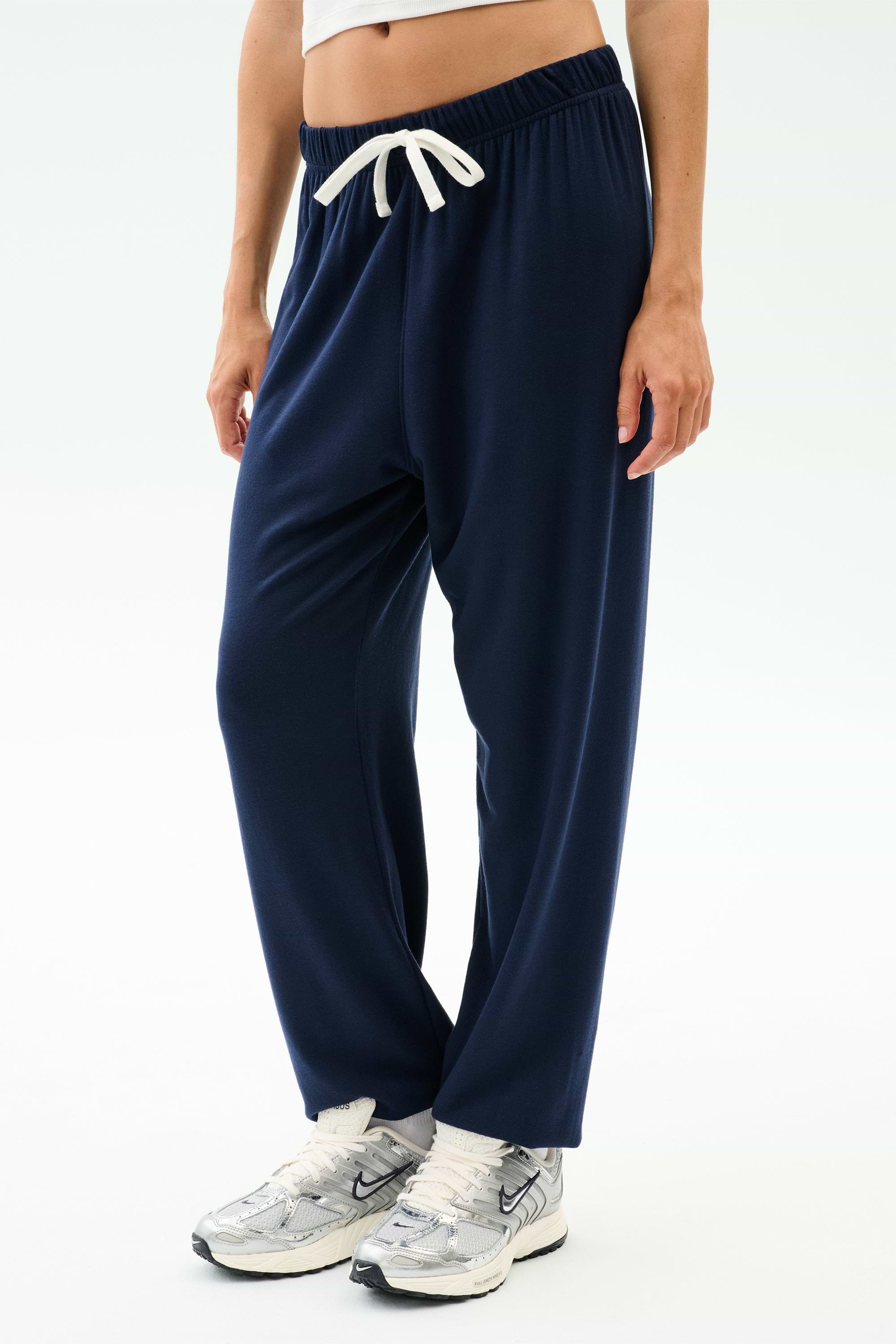 A person wears SPLITS59's Andie Oversized Fleece Sweatpant in Indigo, paired with white sneakers and a cropped white top, standing against a plain white background.