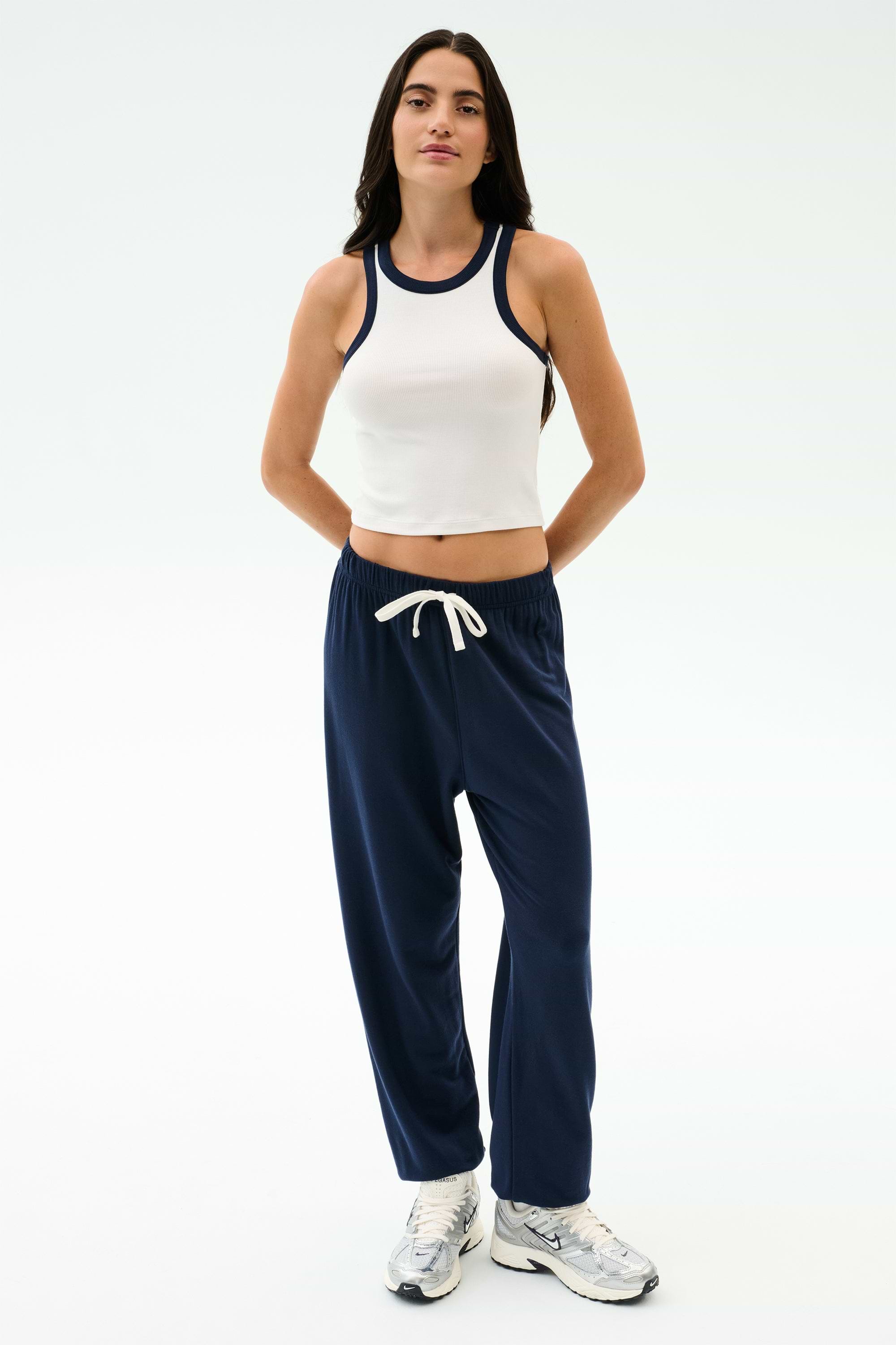 A person wearing a white sleeveless crop top, SPLITS59 Andie Oversized Fleece Sweatpant in Indigo, and white athletic shoes stands against a plain light background.