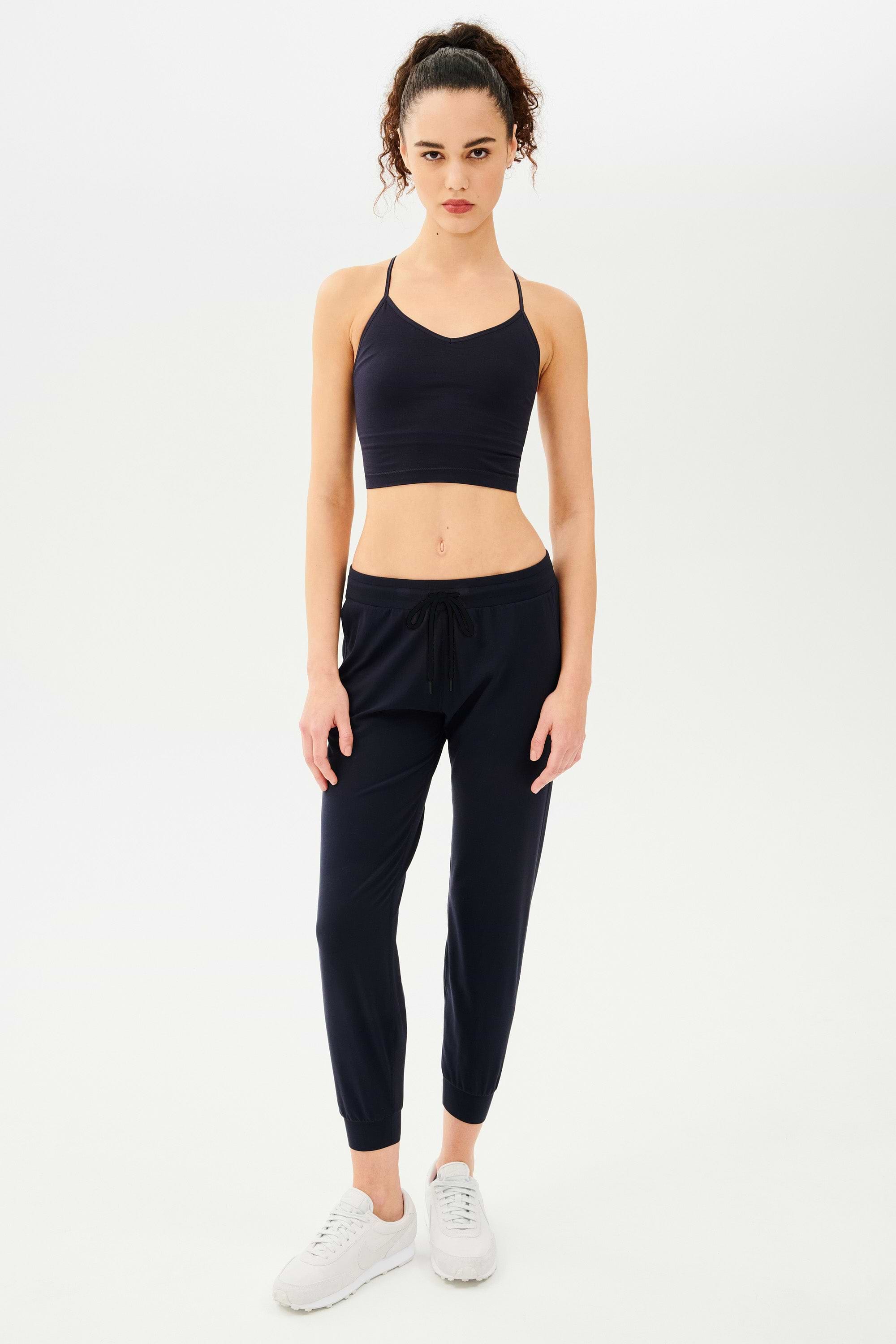Full front view of girl wearing dark blue sweatpants with black tie around waistband and a dark blue sports bra with white shoes