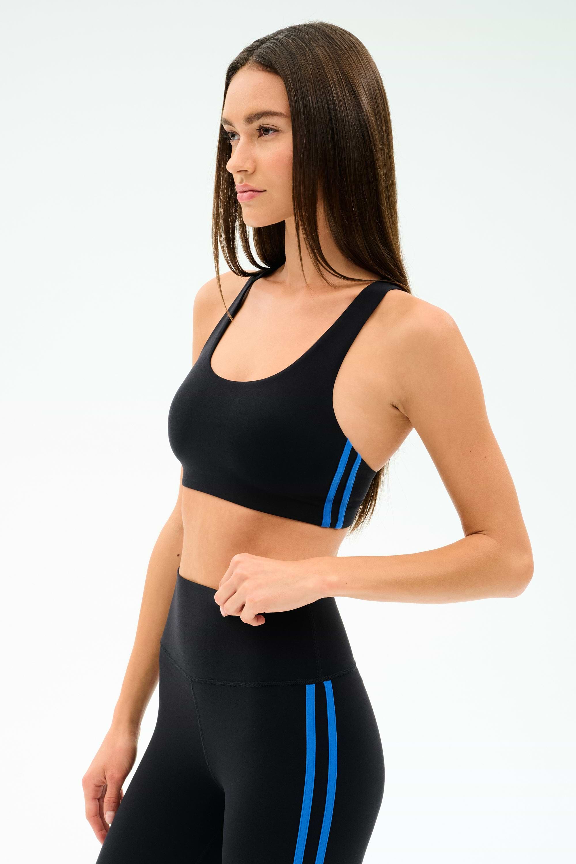 A woman wears the SPLITS59 Ella Airweight Bra in Black/Classic Blue, featuring soft nylon-spandex fabric, paired with high-waisted leggings, standing against a plain white background.