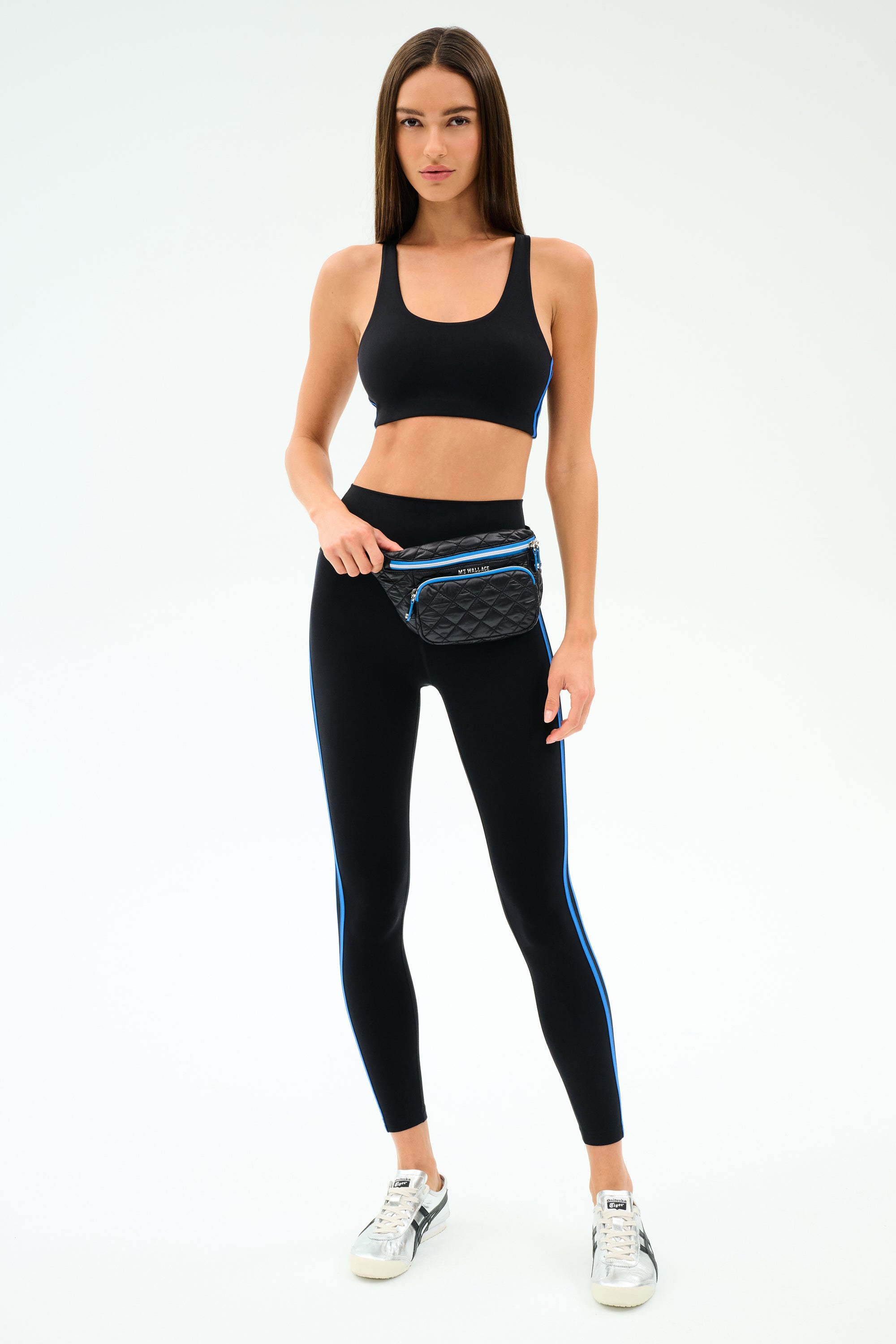 A woman stands against a white background wearing a black sports bra, black leggings with blue stripes, the SPLITS59 MZW x Splits59 Small Metro Sling, and white sneakers.