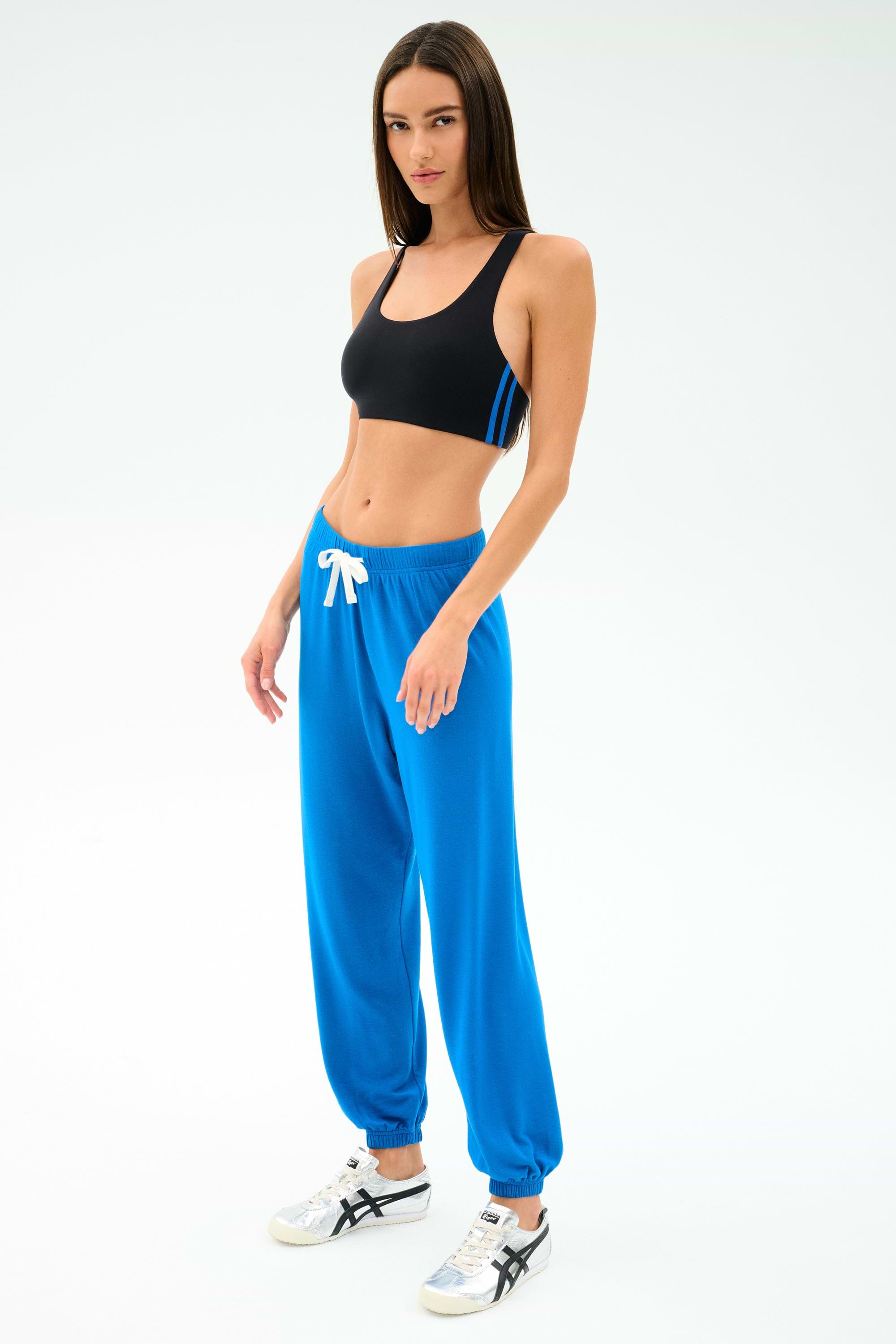A woman in a black spandex sports bra, SPLITS59 Andie Oversized Fleece Sweatpant in Classic Blue with a white drawstring, and white sneakers stands against a plain light background.