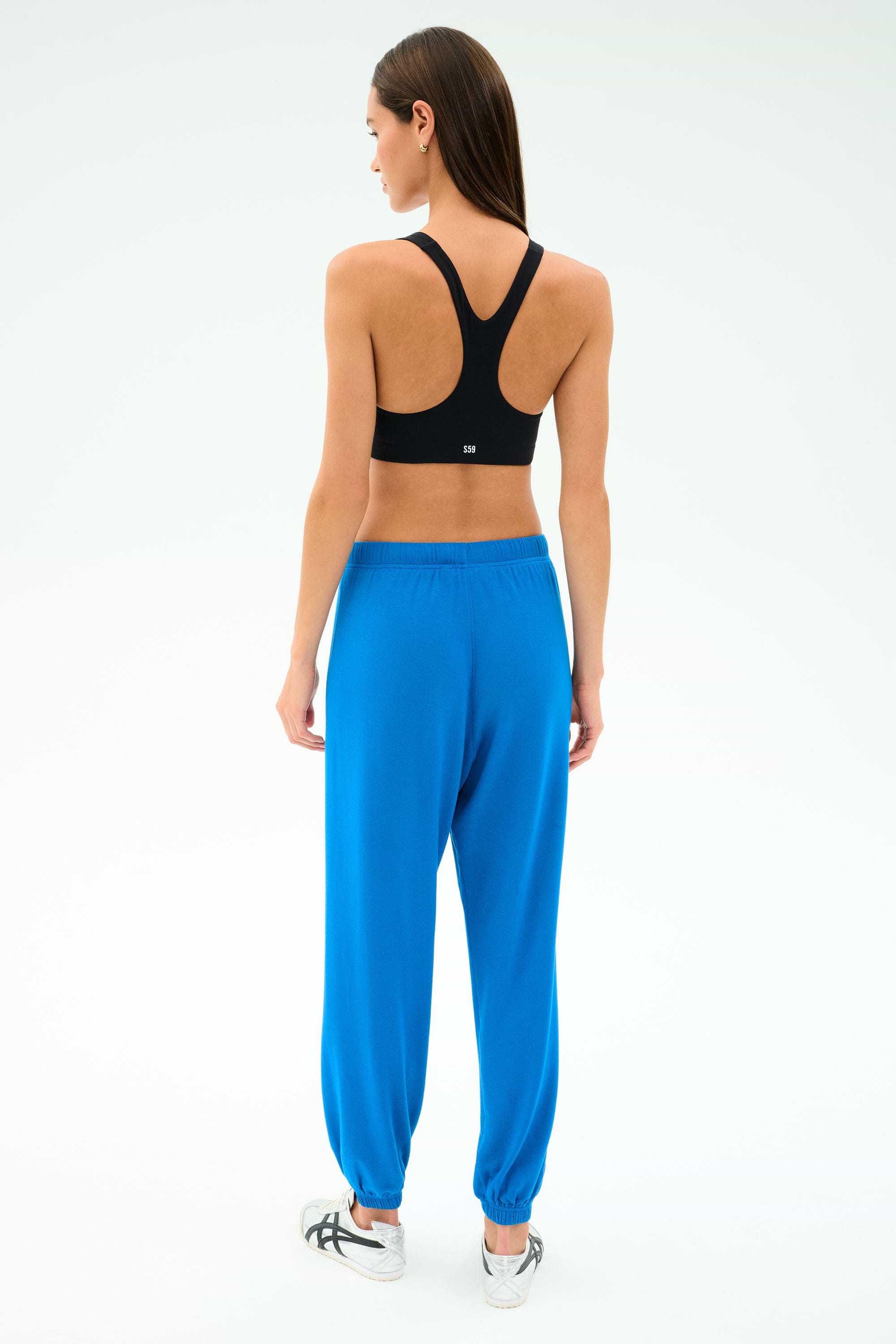 A person stands facing away, wearing a black spandex sports bra, white sneakers, and SPLITS59's Andie Oversized Fleece Sweatpant in Classic Blue against a plain light background.