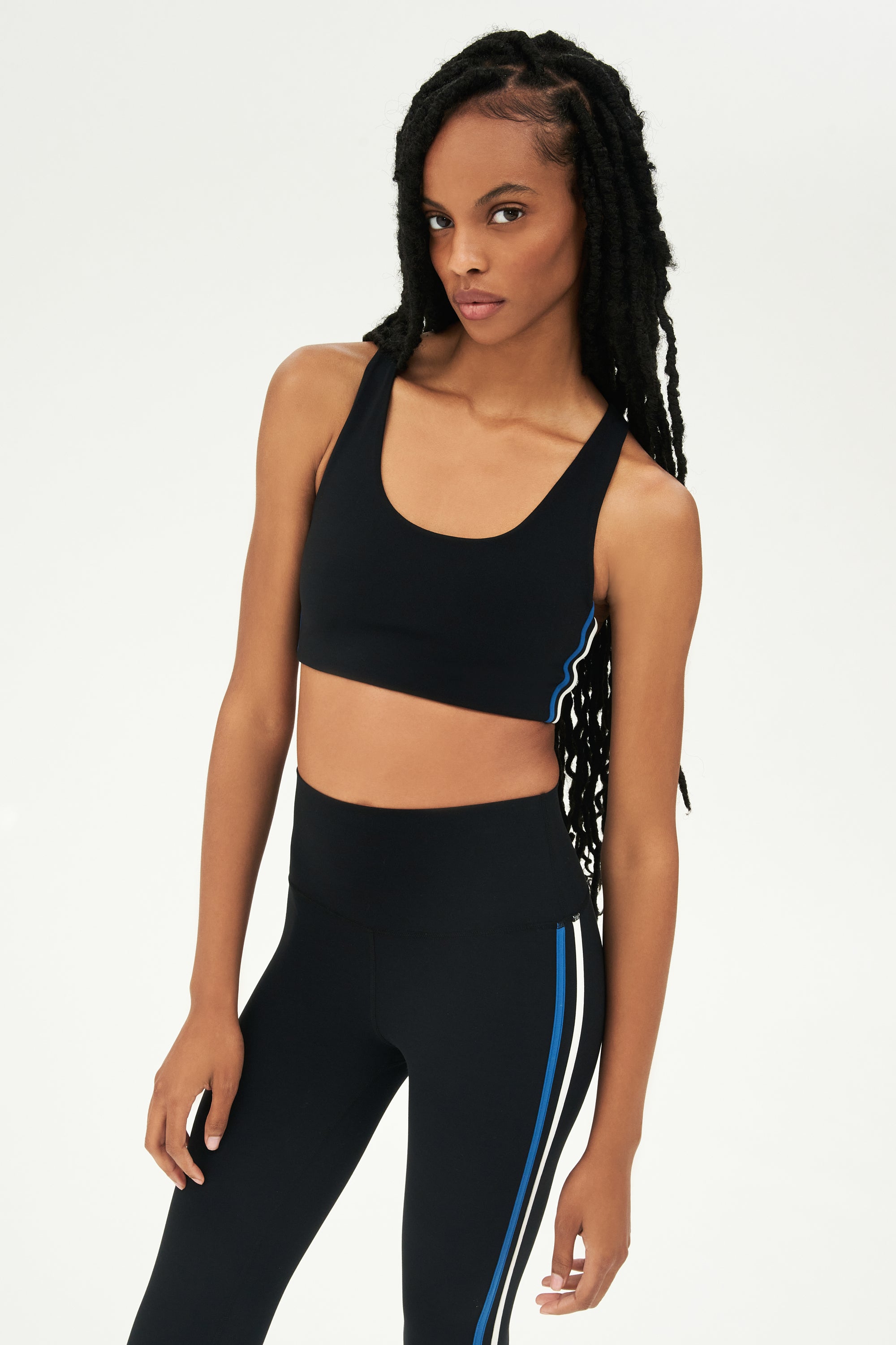 Side view of girl wearing black sports bra with thin white and blue stripes down the side and black leggings 