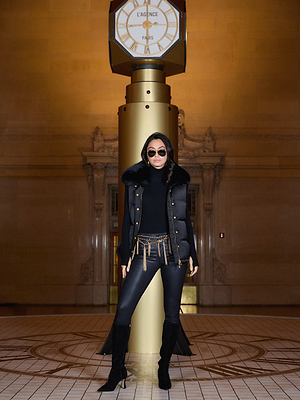 A woman in black sunglasses, a fur-trimmed vest with statement buttons, skinny pants, heeled boots, and the Colette Cropped Turtleneck stands confidently before a large gold clock inside an elegant building.