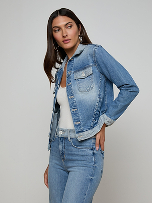 A woman with long dark hair models the Maven Jean Jacket in a light wash over a white top and matching stretch denim jeans, posing with one hand on her hip against a plain light background.