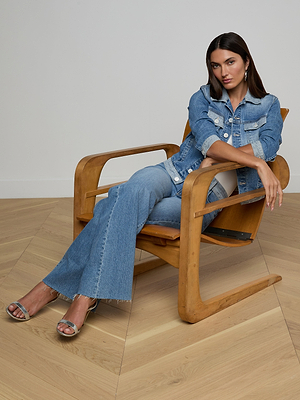 A woman in the Maven Jean Jacket and flared jeans sits confidently in a modern wooden chair on a light herringbone floor, with one arm on the armrest and the other crossed, looking at the camera.