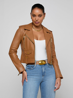 A woman poses against a plain gray background wearing the Blakely Leather Biker Jacket in light brown lambskin over a white top, blue jeans, and a wide belt with a gold buckle. She accessorizes with gold earrings, a necklace, and has her hair pulled back.