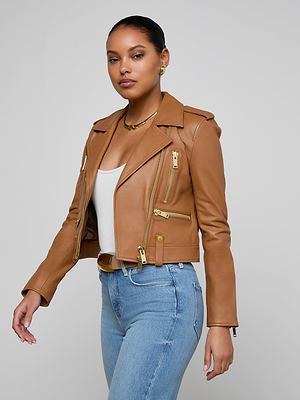 A woman stands confidently against a light gray background, wearing the Blakely Leather Biker Jacket in tan lambskin over a white top and blue jeans, looking directly at the camera.