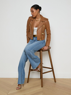 A woman sits on a wooden stool against a white wall, wearing the Blakely Leather Biker Jacket over a white top, paired with blue flared jeans and heeled sandals. She looks down to her left, hands resting on the stool.