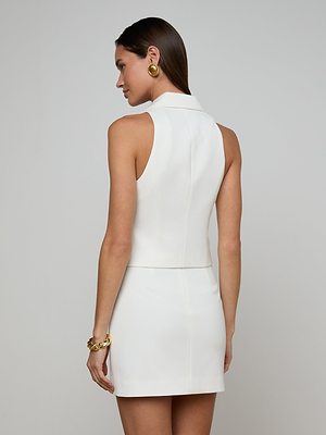 A woman with long brown hair wears the Tomi Vest—a sleeveless white dress with a high collar and satin lapel—styled with gold earrings and a chunky gold bracelet, posing against a plain background.