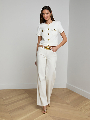 A woman stands indoors on a wooden floor, wearing the tailored Nawa Shirt Jacket in white with gold buttons, wide-leg white pants, a white belt with a gold buckle, gold earrings, and heeled sandals.