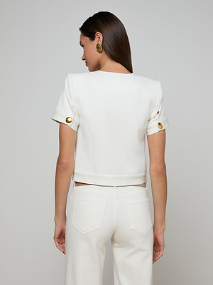 A woman with straight brown hair is seen from behind wearing the Nawa Shirt Jacket—a tailored, short sleeve white top with gold sleeve buttons—paired with white pants, set against a plain light background.