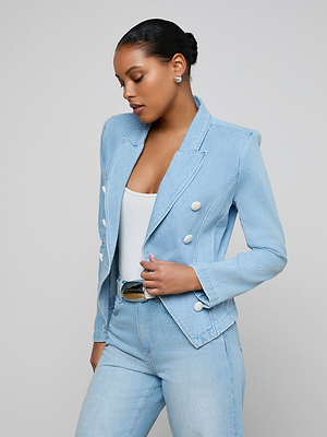 A woman wears the Emerson Structured Blazer in light blue denim with large white buttons, paired with a white top and matching denim pants, standing against a plain light gray background and looking down with her hands near her waist.