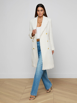 A woman with long brown hair and gold accessories stands on a wooden floor against a white wall, wearing the Cooper Tweed Trench Coat—a long white tweed jacket with frayed trims—over a white top and blue jeans.