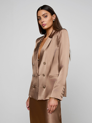 A woman with long brown hair wears the shiny, light brown Sena Silk Blazer over a matching dress. She stands against a plain light background, looking at the camera with a neutral expression.
