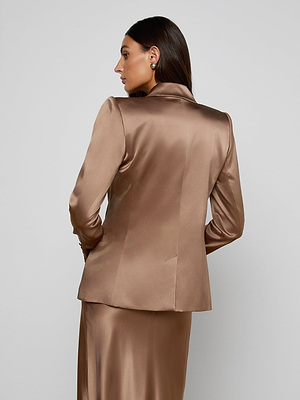 A woman with long brown hair models the Sena Silk Blazer in shiny bronze with a matching skirt, standing against a plain light gray background and glancing over her right shoulder.