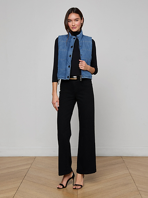A woman smiles at the camera indoors on a wooden floor, wearing the indigo Rola Puffer Vest over a black long-sleeve top, with black wide-leg pants and black heeled sandals.