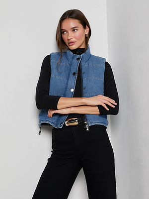 A woman with long brown hair wears the cropped, sleeveless indigo Rola Puffer Vest over a black long-sleeve top and pants, standing with arms crossed against a white wall and looking to the side.