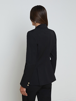A person with long brown hair, wearing the L’AGENCE Chamberlain Blazer and matching black pants, stands facing away from the camera against a plain white wall. Gold accent buttons detail the blazer's sleeves.