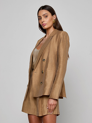 A woman with long brown hair wears a gold Colin Linen-Blend Blazer over a tan top, paired with matching shorts. She stands against a plain light gray background, looking confidently at the camera.