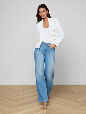 A woman stands indoors with one hand in her pocket, wearing the Brooke Blazer—an open-front white blazer with gold buttons—over a white top, light blue wide-leg jeans, and white heels. She has long hair and poses against a plain wall on wooden flooring.
