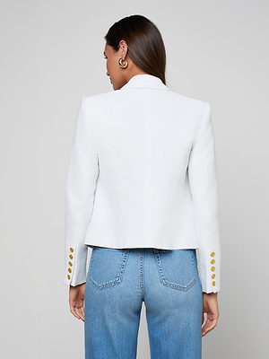 A woman with long brown hair is seen from behind wearing the Brooke Blazer—an open-front white blazer with gold sleeve buttons—and blue jeans, against a light gray background.