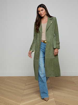 Celina models the Celina Suede Trench Coat in olive green over a beige top, blue jeans, and tan pointed heels. She poses indoors by a light wall and wood floor, her long brown hair flowing down.