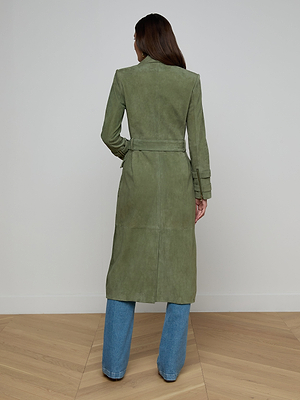 Seen from behind, a person with long brown hair wears the Celina Suede Trench Coat and blue jeans, standing on a light wood floor against a plain light wall—a timeless wardrobe essential.