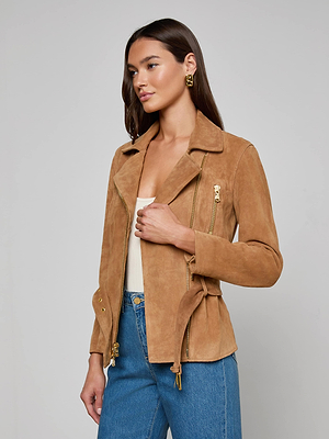 A woman with long brown hair wears the Dinah Suede Biker Jacket with gold zips over a white top and blue jeans against a light gray background. She accessorizes with gold earrings and holds the jacket lapel.
