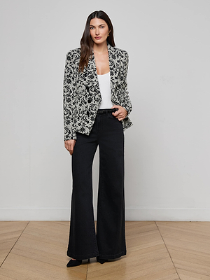 A woman with long brown hair stands against a plain wall, wearing the Bethany Flocked Tweed Blazer over a white top, paired with black wide-leg pants and pointed-toe shoes. She poses relaxedly with one hand in her pocket.