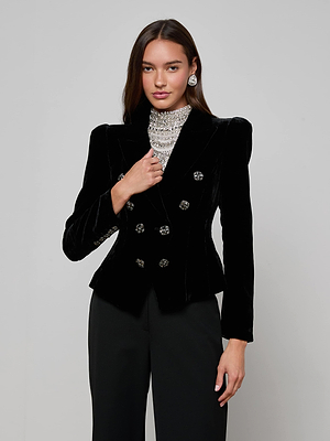 A woman wears the L'AGENCE Bethany Velvet Blazer—a black double-breasted style with crystal-embellished buttons—paired with black pants, large statement earrings, and a jeweled high-neck top against a plain gray background.