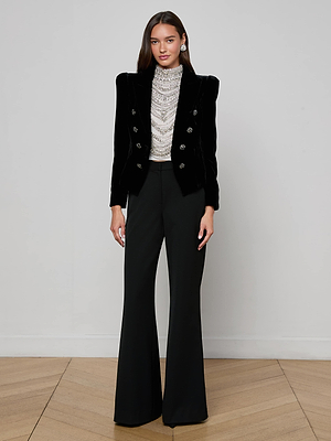 A woman with long, loose hair stands against a plain white wall, wearing the L'AGENCE Bethany Velvet Blazer with crystal buttons over a high-neck silver beaded top and high-waisted black wide-leg trousers.