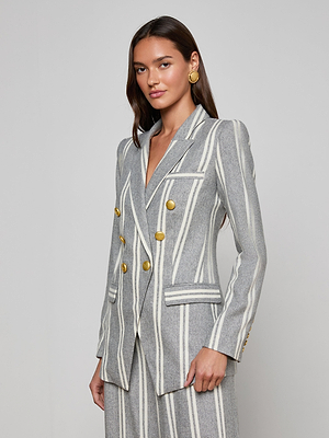 A woman stands against a plain background wearing the Kendi Longline Blazer in gray with white vertical stripes, gold buttons, and matching pants. She has long brown hair and accessorizes with large round gold earrings.