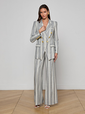 A woman poses against a plain light gray wall in the Kendi Longline Blazer, a gray Italian wool blend suit with white vertical stripes, gold buttons, and wide-leg trousers. She wears gold earrings, white heeled sandals, and has long brown hair.