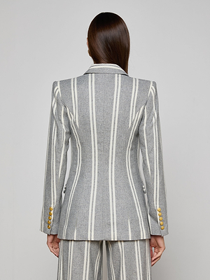 A person with long brown hair is shown from the back, wearing the Kendi Longline Blazer—a gray Italian wool blend suit with vertical white stripes, gold sleeve buttons, and matching fitted pants—against a plain background.