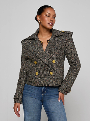 A woman stands against a plain gray background wearing the Xoey Cropped Tweed Trench—a double-breasted, oversized Italian wool-blend jacket with gold buttons—paired with blue jeans, gold hoop earrings, and her dark hair pulled back.