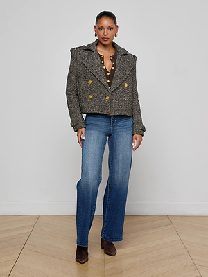 A woman stands against a plain wall wearing the Xoey Cropped Tweed Trench, a brown polka dot blouse, blue jeans, and brown ankle boots on a wooden floor.