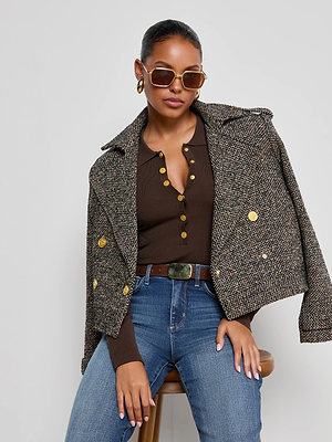 A stylish woman poses on a stool, wearing sunglasses, gold hoop earrings, blue jeans, a brown buttoned top, and the Xoey Cropped Tweed Trench draped over her shoulders against a plain background.