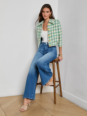 A woman with long brown hair and gold hoop earrings sits on a wooden stool against a white wall, wearing the Jia Houndstooth Tweed Jacket in jade green, a white top, blue wide-leg jeans, and beige heels.