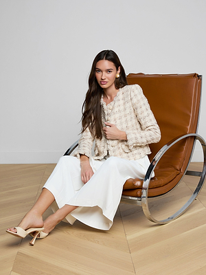 A woman with long brown hair sits on a modern brown leather and chrome chair, wearing the Zell Houndstooth Tweed Jacket, white wide-leg pants, and beige heels against a neutral background with a wooden floor.